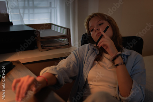 Young female student feeling bored and tired during late night study, procrastinating on homework or school project in a dimly lit home office