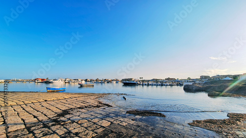 Panoramic view of the traditional stone boat slipway in the old seaside village of Marzamemi, Sicily, Italy, showcasing fishing boats moored in a calm blue harbor under a sunny summer sky.
