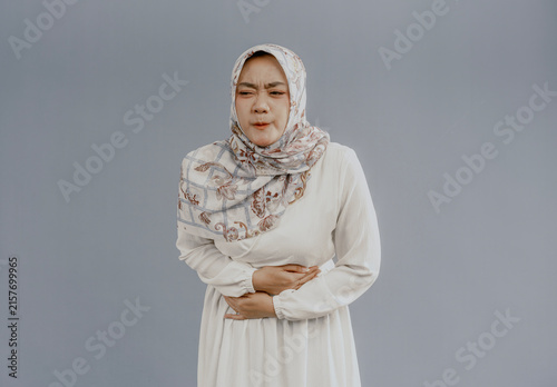Asian muslim woman in hijab experiencing stomach ache, bending over and holding hands on stomach, discomfort due to menstrual cramps, closing eyes, isolated on white background.