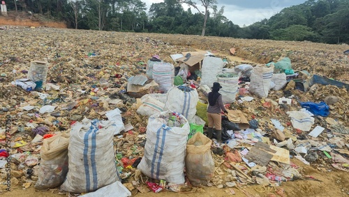 Картината върху платно KENDARI, INDONESIA - JULY 24, 2026: A scavenger woman stands among large sacks filled with sorted recyclable plastic waste collected manually at the Puuwatu landfill site
