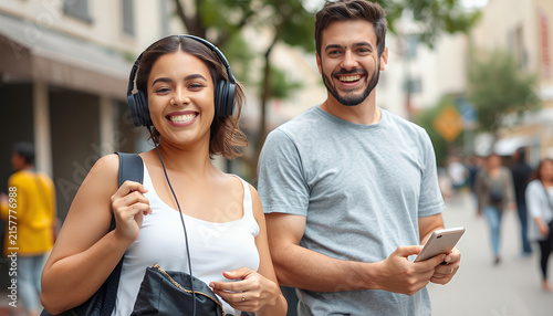 happy couple walking outdoors on a sunny city street, woman wearing white sleeveless top with headphones and backpack, man