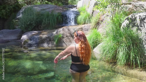 Rear view of a red-haired woman in a natural river pool, gazing at a small waterfall. Sierra de Gredos, Spain