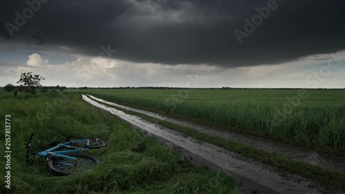 blue bike on the edge of a wet dirt track through a lush green wheat field under a dramatic dark stormy sky during rainfall