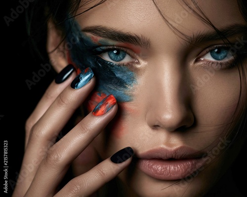Extreme close-up of a woman with blue eyes and blue red face paint touching her cheek with black and colorful manicured nails Expression of artistic beauty