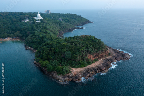 Bild auf Leinwand Aerial perspective of a densely forested rocky headland jutting out into the calm blue sea, with a majestic white peace pagoda visible in the background