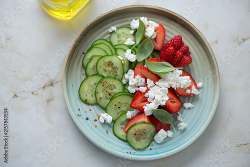 Salad with fresh strawberries, cucumber and ricotta on a turquoise plate, horizontal shot on a light-beige marble background, high angle view