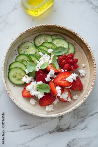 Beige bowl with strawberry, cucumber and ricotta salad, vertical shot on a white marble background, high angle view