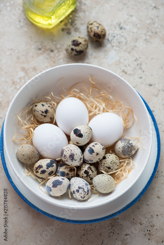 White bowl with uncooked quail and chicken eggs, vertical shot on a beige granite background, elevated view, selective focus