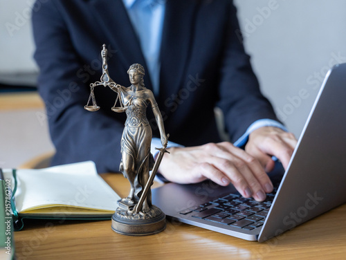 Lawyer in dark suit typing on silver laptop. Bronze Lady Justice figurine stands on wooden desk. Open notebook and pen nearby. Modern digital legal practice with traditional justice symbol in office.
