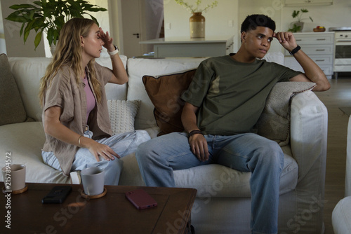 Canvas Print Diverse couple sitting apart on light-colored sofa in living room, smartphones a