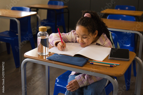 African American child leaning over notebook at desk, writing with red pencil, pink striped top