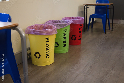Three recycling bins sitting against cream wall on wood-look floor near student desks and chairs