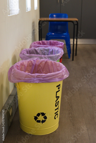 Yellow recycling bin is sitting along left wall in corridor, showing PLASTIC text and purple liner