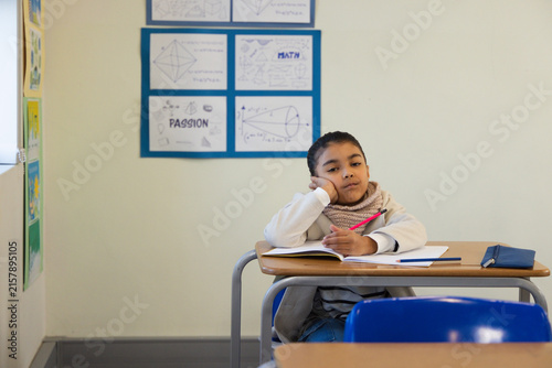 African American school-age girl sitting at classroom desk writing in notebook with pink pencil