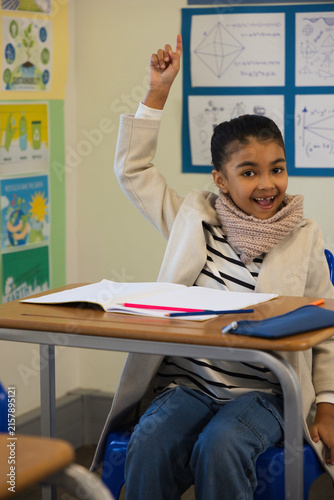 African American child raising hand, sitting at desk in classroom with notebook and pens