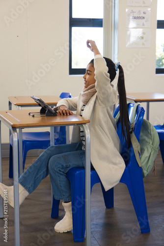 Tablet is sitting propped on wooden desk in classroom with blue chair and backpack near windows