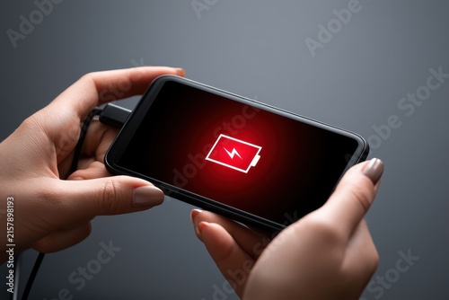 Woman hands plugging a charger in smart phone close-up, dark background. Smartphone with red warning low battery icon on screen charging power. Mobile phone charge. Cellphone is charged via USB cable