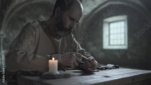 Bearded medieval man locked in iron shackles cutting a quill pen with a small knife, while sitting at a rough wooden table next to a burning candle inside a cold dungeon room