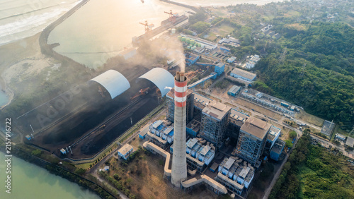 Aerial view of a coal-fired power plant with a tall chimney emitting smoke, coal storage piles under arched covers, and industrial infrastructure along the coast in Indonesia.