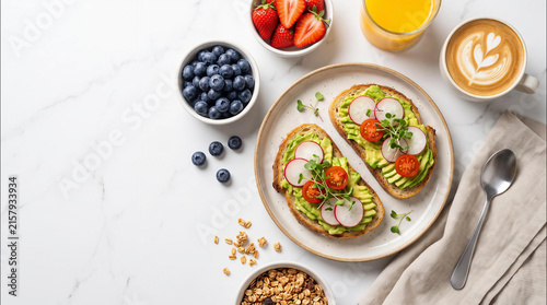 Delicious avocado toast topped with fresh radishes and tomatoes served on white marble surface with berries granola coffee and juice