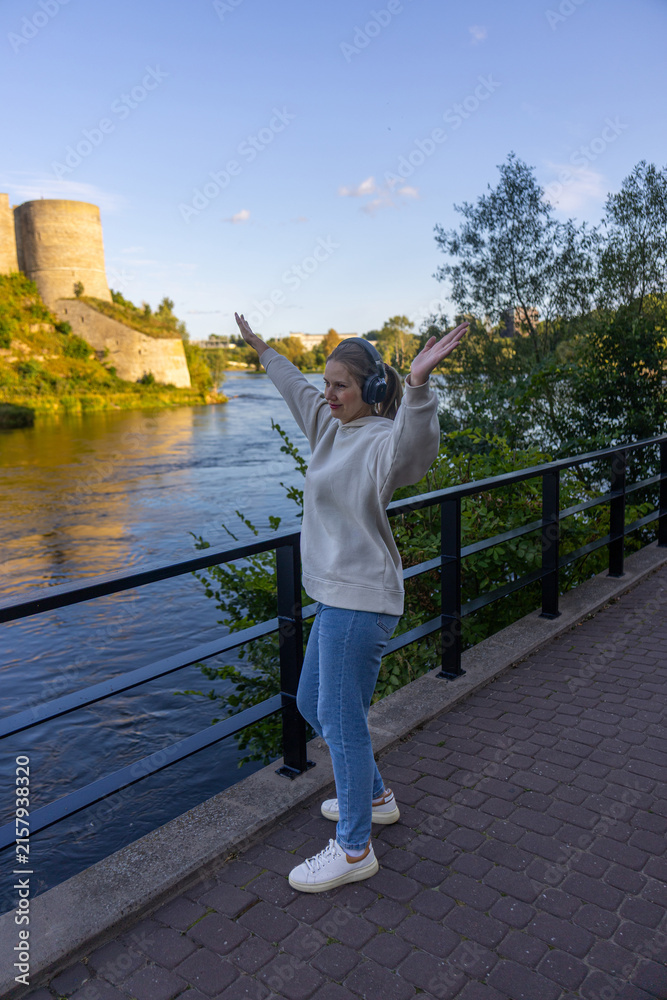 Fototapeta premium Woman with headphones dancing joyfully on riverside walkway by fortress