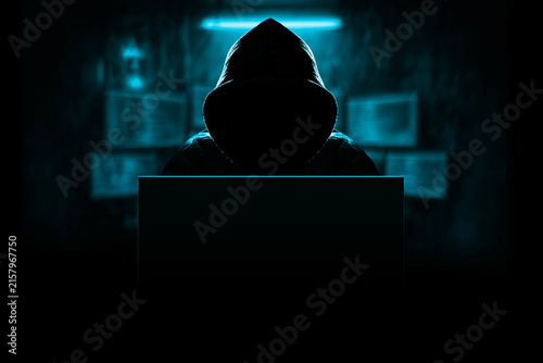 Konstfotografi Faceless person in hoodie behind laptop surrounded by screens, symbolizing cyberattack, data breach risk and digital security threats