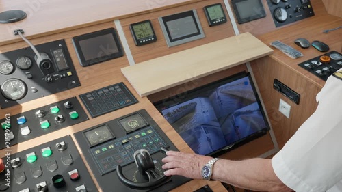 A ship captain navigates a commercial vessel using the modern bridge control console and joystick. Digital screens display live navigation views during precise boat maneuvering in a harbor.