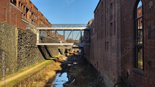 A cityscape featuring a metal bridge crossing historic brick buildings over a shallow canal in Georgetown. A clear sunny day highlights the strict geometry and texture of the walls.