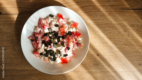 A white plate features a fresh salad with diced tomatoes, creamy base, and pumpkin seeds; sunlight stripes fall across the plate and wooden table, emphasizing textures.