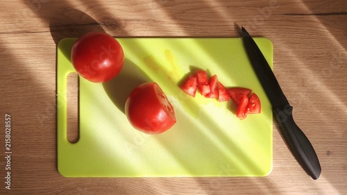 Two whole red tomatoes and diced pieces on a bright green cutting board; black knife placed diagonally; sunlight accentuates textures on the wooden tabletop.