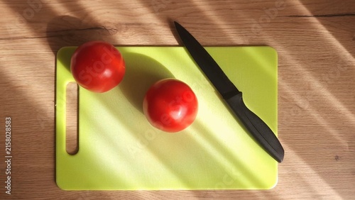 Two ripe red tomatoes and a sleek black knife rest on a bright lime-green cutting board; warm sunlight casts diagonal stripes across the wooden surface.