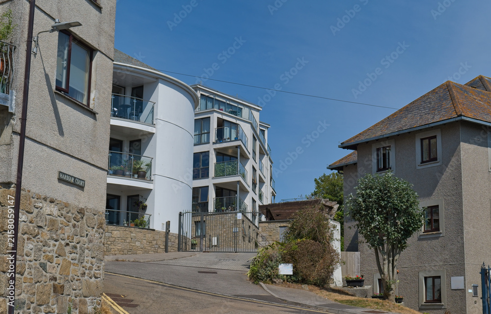 Fototapeta premium A sunny street scene showing modern buildings on the left, traditional homes on the right, and a paved road leading uphill - Penzance, Cornwall, UK