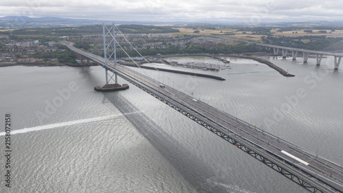 Aerial view of the Forth Road Bridge spanning the Firth of Forth with the Queensferry Crossing in the background, featuring steel suspension cables in South Queensferry, United Kingdom.