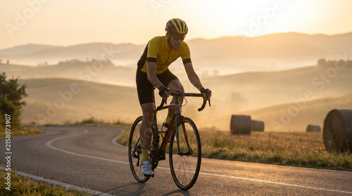 Cyclist riding a road bike through rolling hills at sunrise with hay bales