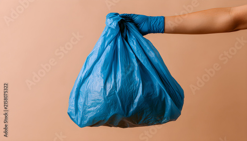 Hand wearing blue nitrile glove holds plastic bag filled with household waste against beige background. Person carries full sack of garbage for disposal. Concept of recycling sustainability