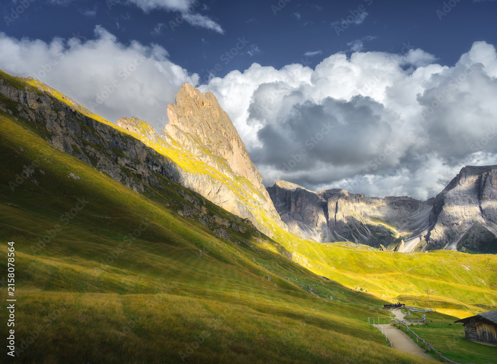Naklejka premium Seceda mountain peak with green grass meadow slope under dramatic cloudy sky at sunset in Dolomites, Italy. Alpine autumn landscape with rocky cliffs, sunbeams, wooden hut and hiking path. Travel