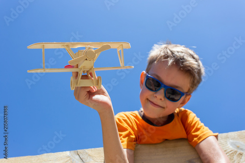 A fair-haired boy in sunglasses stands on a fence and holds a wooden airplane in his hand against the sky.