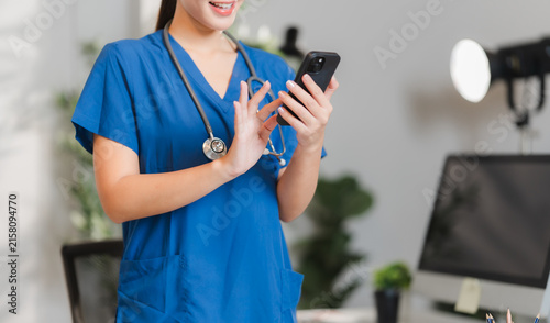 Obraz na płótnie Healthcare Professional with Smartphone: A smiling healthcare worker in blue scrubs, wearing a stethoscope around her neck, is engrossed in her smartphone