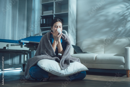 Canvastavla Ailing Woman's Comfort: A young woman, wrapped in a warm blanket and seated on cushions, blows her nose into a tissue while feeling unwell at home, highlighting illness and recovery
