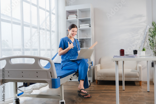 Konstfotografi Virtual Healthcare Consultation: A friendly nurse in blue scrubs engages in a telehealth consultation via laptop, seated on a hospital bed in a well-lit room