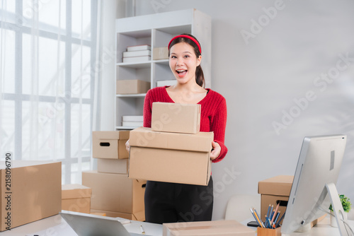 Young Woman Entrepreneur Holding Package: A cheerful young woman, an aspiring entrepreneur, beams with excitement as she holds a stack of cardboard packages, ready for shipping, in her home office.