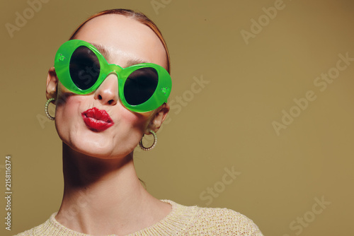 Принти на полотні Young woman with bright red lipstick wearing large green sunglasses and hoop earrings poses with puckered lips on beige background