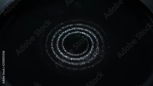 Tiny particles form concentric circles on a dark surface. The shimmering pattern creates a mesmerizing visual effect.
