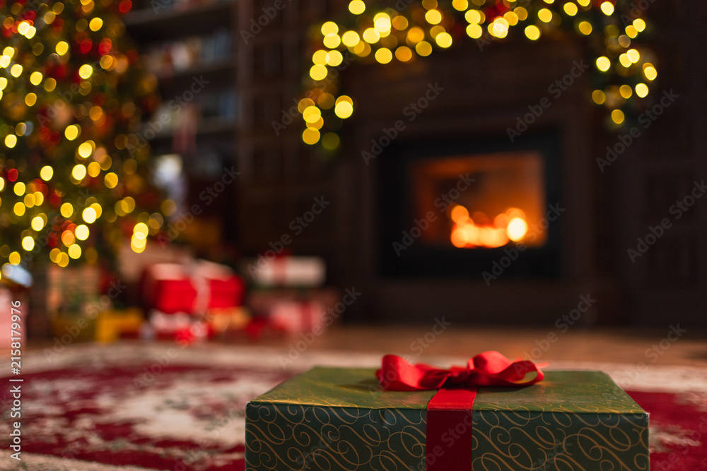 Fototapeta premium Green gift box in classical Christmas decorated interior living room library with fireplace. Christmas gift near fir tree on Christmas eve. Modern classic style interior design apartment. Christmas