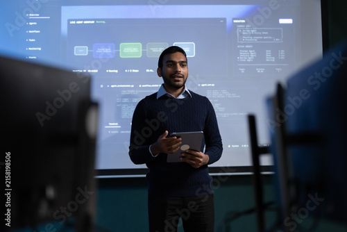 Young Indian male professional holds a tablet while presenting a software workflow to coworkers in a meeting room, with computer screens and listeners in the foreground.