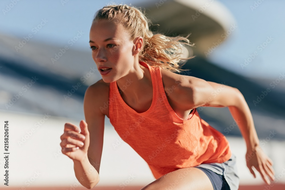 Fototapeta premium Female runner accelerating from start on athletics track in orange tank top for training banner and sports background