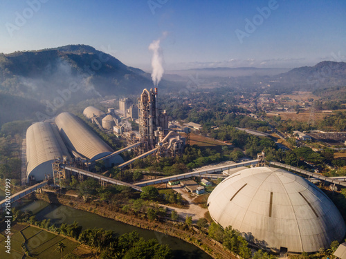 Aerial view of a large industrial cement factory complex with a tall chimney emitting smoke, large storage domes, and conveyor belts, situated amidst lush green hills in Indonesia.