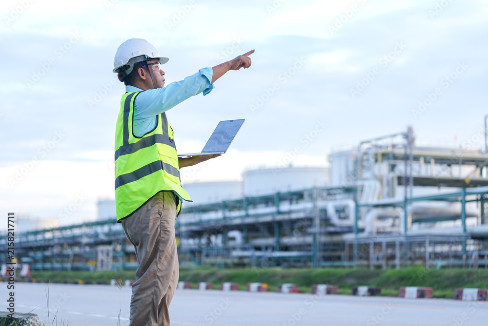 Fototapeta premium Engineer safety vest hard hat laptop pointing industrial plant pipeline inspection, industrial worker monitoring refinery facility for safety compliance and maintenance planning outdoors
