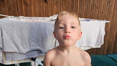 Blonde boy with a funny puckered-lip expression poses shirtless in front of a drying rack with clothes; rustic wooden wall and dark green fabric add a cozy domestic feel