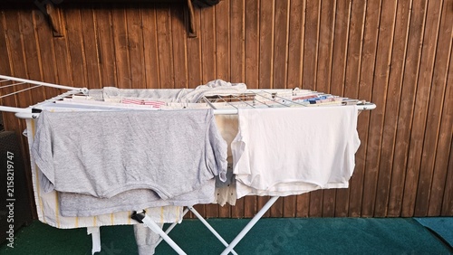 A white folding clothes drying rack holds grey and white garments; small items are clipped on the upper tier. The backdrop features vertical wooden panels and a dark green floor